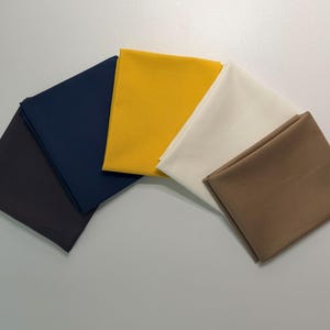 Puede incluir: Vista aérea de cinco cuadrados de tela doblados en forma de abanico. Los colores incluyen gris oscuro, azul marino, amarillo dorado, blanco roto y marrón. Las telas parecen lisas, probablemente de algodón o material similar.