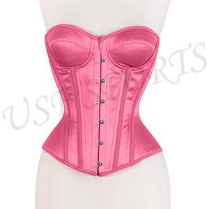 May include: A pink satin corset displayed on a mannequin. The corset features a structured design with vertical boning, a sweetheart neckline, and a row of silver-toned buttons down the front. The garment is a vibrant shade of pink.