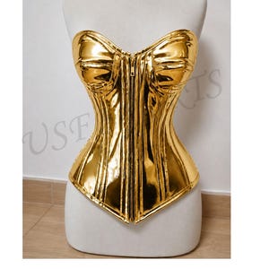 May include: A gold-coloured corset with a central zip fastening, displayed on a white mannequin. The material has a metallic sheen, possibly faux leather. The corset has a structured, fitted design, with a bustier-style top.
