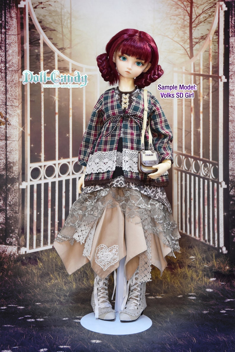May include: A doll wearing a plaid shirt with lace trim, a beige skirt with lace overlay, and white lace-up boots. The doll has long, dark red hair and blue eyes. The doll is standing in front of a metal gate with a sign that reads "Doll-Candy www.doll-candy.com". The sign also says "Sample Model: Volks SD Girl".