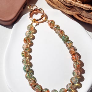 May include: A beaded phone strap with a gold-tone ring and clasp. The beads are clear with orange and green accents. The strap is on a white plate with a brown phone case in the background.