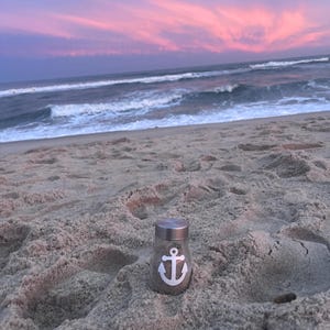 May include: A small glass jar with a silver lid and a white anchor design, filled with sand, sits on a sandy beach. The background features a vibrant sunset with pink and purple hues over the ocean.
