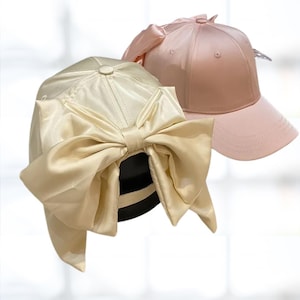 May include: Two satin baseball caps with large bows on the back. One cap is cream coloured and the other is pink.