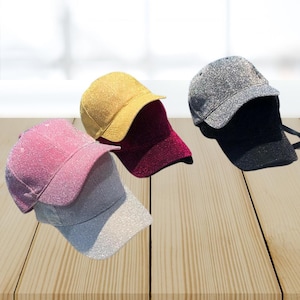 May include: Four glitter baseball caps in pink, silver, gold, and burgundy. The caps are arranged on a wooden surface, with the pink and silver caps in the foreground. The caps have a classic six-panel design with a curved brim.