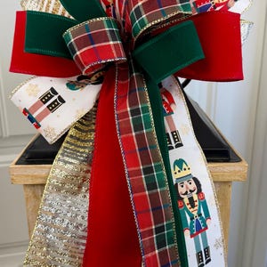 May include: A festive Christmas bow featuring a variety of ribbons. The bow includes red velvet, green, gold, and plaid ribbons. Some ribbons display nutcracker figures. This decorative bow is perfect for holiday decorating.