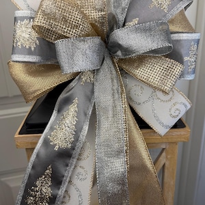 May include: A decorative Christmas bow featuring multiple ribbons in gold, silver, gray, and white. The bow has a layered design with gold glitter accents and a Christmas tree pattern on the gray ribbon. The bow is placed on a wooden lantern.