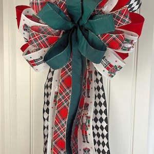 May include: A festive Christmas bow with a large green bow in the center. The bow features red, white, and green ribbons with plaid, diamond, and nutcracker designs. The bow is attached to a white door.