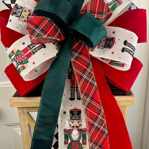 May include: A festive Christmas bow featuring red, green, and white ribbons. The bow has a plaid pattern and a ribbon with a nutcracker soldier design. The bow is a decorative accent for the holiday season.