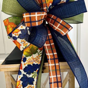 May include: A decorative bow featuring a mix of ribbons in fall colors. The bow includes ribbons with orange and white plaid, navy blue, and green. Some ribbons have a pattern of autumn leaves and berries. The bow is likely for seasonal decor.