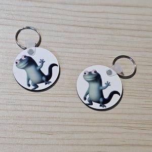 May include: Two circular keyrings with a cartoon lizard character in blue and grey. The lizard is smiling and waving on a white background. Each keyring has a silver metal ring attached.