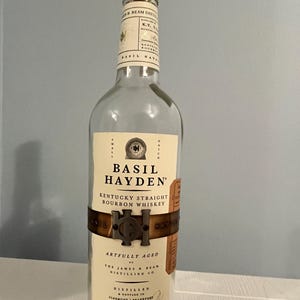 Puede incluir: Una botella de vidrio transparente de Basil Hayden's Kentucky Straight Bourbon Whiskey. La botella tiene una etiqueta marrón con letras doradas y una banda metálica alrededor del medio. La etiqueta dice "Basil Hayden's Kentucky Straight Bourbon Whiskey" y "Artfully Aged by The James B. Beam Distilling Co.". La botella es de 750 ml y 100 proof.