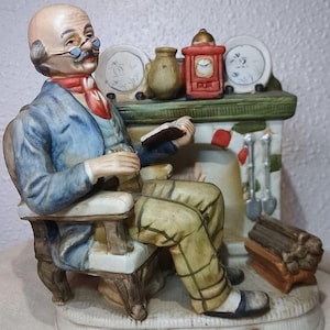 Peut inclure: Figurine en céramique d'un vieil homme assis dans un fauteuil, lisant un livre. Il porte une veste bleue, un pantalon à motifs et des lunettes. Une cheminée avec une horloge, des assiettes et des outils est derrière lui. La figurine est sur une base décorative.