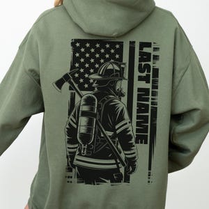 May include: Khaki green hoodie with a graphic of a firefighter and an American flag design, with the text "LAST NAME". The design includes a firefighter with an axe and oxygen tank. The hoodie has a hood and long sleeves.