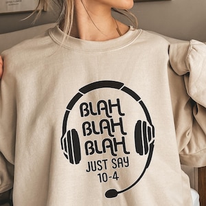 May include: Beige sweatshirt with a black graphic of a headset and the words "BLAH BLAH BLAH JUST SAY 10-4". The sweatshirt is a neutral color and has long sleeves.