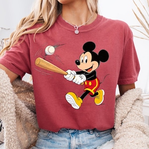 May include: A maroon t-shirt with a cartoon image of Mickey Mouse playing baseball. Mickey Mouse is depicted swinging a bat at a baseball. He wears yellow shoes, red shorts, white gloves, and a black shirt. The baseball is white.