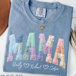 May include: Blue jean Comfort Colors t-shirt with the word "MAMA" in patchwork letters. The letters are in various colors and patterns. Below the word "MAMA" are the names "Emily", "Robert", and "Sofia". A gold necklace is draped across the shirt.