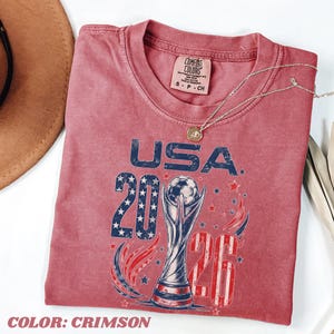 K&ouml;nnte beinhalten: Ein karmesinrotes T-Shirt mit einer Grafik, die den Text USA 2026, einen Fu&szlig;ballpokal sowie Sterne- und Streifenmuster zeigt. Das Shirt hat ein Comfort Colors-Etikett am Nacken und einen Rundhalsausschnitt. Eine d&uuml;nne goldfarbene Kette liegt auf dem Stoff.