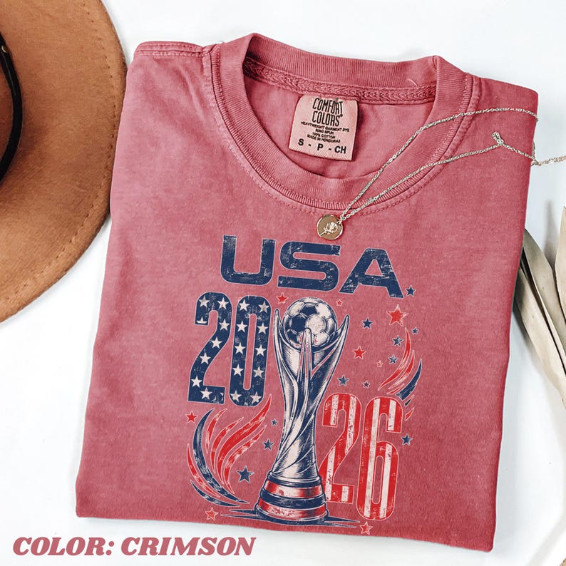 K&ouml;nnte beinhalten: Ein karmesinrotes T-Shirt mit einer Grafik, die den Text USA 2026, einen Fu&szlig;ballpokal sowie Sterne- und Streifenmuster zeigt. Das Shirt hat ein Comfort Colors-Etikett am Nacken und einen Rundhalsausschnitt. Eine d&uuml;nne goldfarbene Kette liegt auf dem Stoff.