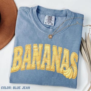 May include: Blue jean coloured t-shirt with the word "BANANAS" in yellow, patchwork-style lettering. A bunch of bananas is on the right side of the word. A gold necklace with a pendant is on the shirt.