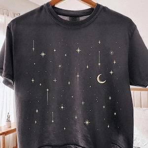 May include: A charcoal gray short-sleeve t-shirt featuring a celestial graphic print. The design consists of various gold-colored stars, sparkling points of light, and a crescent moon scattered across the front of the garment.