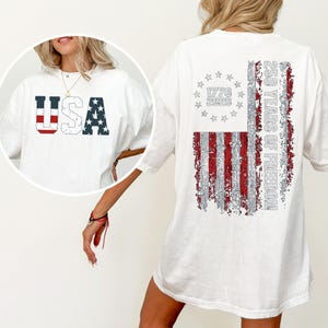 May include: A white t-shirt featuring a distressed graphic on the front that reads USA with red stripes and blue stars. The back displays a large distressed American flag design with a circle of stars, the text 1776 2026 and 250 YEARS OF FREEDOM.