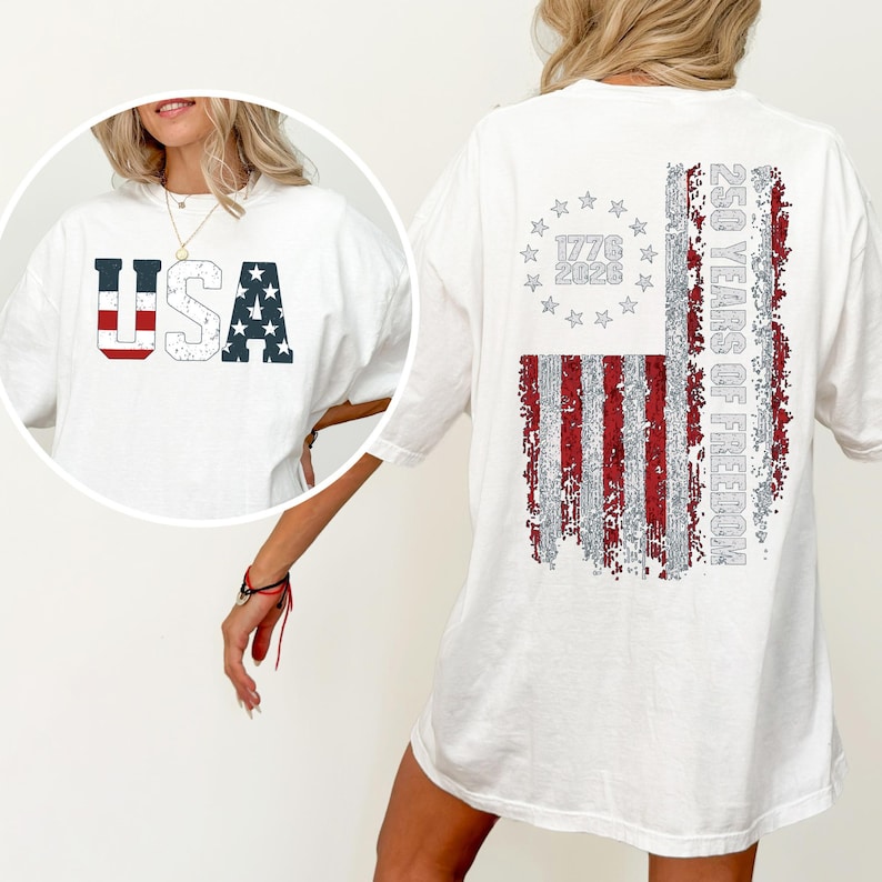 May include: A white t-shirt featuring a distressed graphic on the front that reads USA with red stripes and blue stars. The back displays a large distressed American flag design with a circle of stars, the text 1776 2026 and 250 YEARS OF FREEDOM.