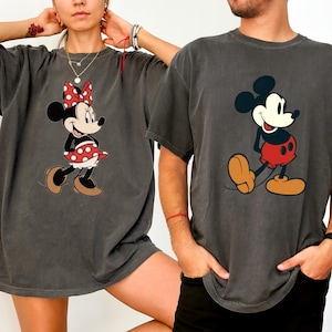 May include: Two charcoal gray t-shirts featuring Disney's Mickey and Minnie Mouse. Minnie is wearing a red polka dot bow and skirt. Mickey is wearing red shorts and yellow shoes. Both shirts have a vintage, faded look.