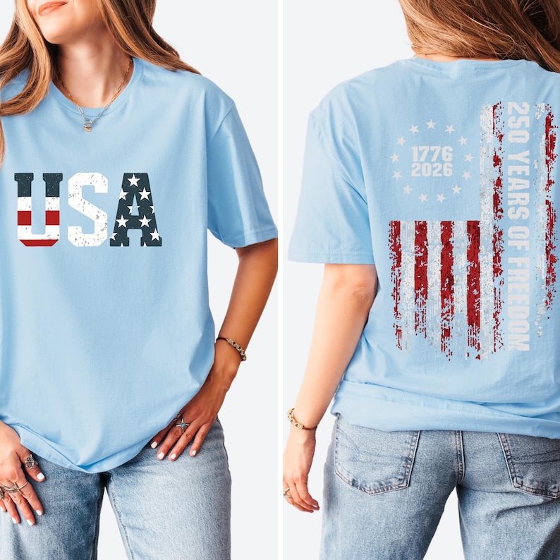 May include: A light blue t-shirt featuring a distressed USA graphic on the front. The back displays a distressed American flag design with a circle of stars, the years 1776 and 2026, and the vertical text 250 YEARS OF FREEDOM in white.
