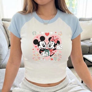 May include: White and light blue raglan crop top featuring a graphic of Mickey and Minnie Mouse inside a pink heart. The cartoon couple is holding a gift, surrounded by hearts and fireworks.