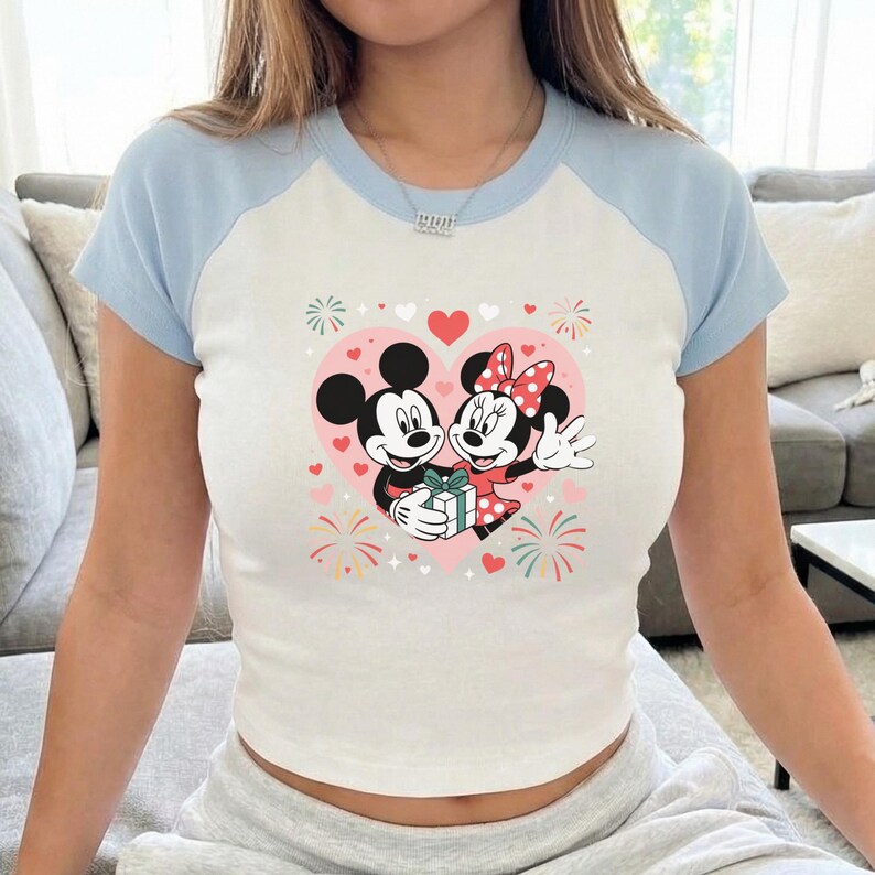 May include: White and light blue raglan crop top featuring a graphic of Mickey and Minnie Mouse inside a pink heart. The cartoon couple is holding a gift, surrounded by hearts and fireworks.