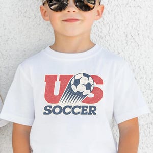 May include: A white short-sleeved t-shirt featuring a graphic design with the red letters US, a black and white soccer ball with motion lines, and the word SOCCER in blue block lettering. The graphic has a distressed, vintage-style texture.