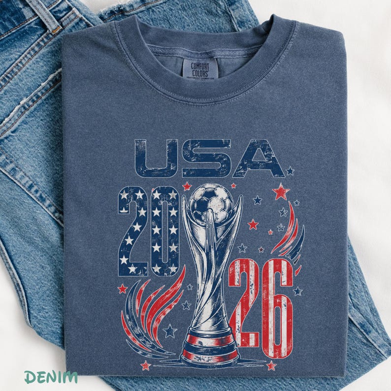 K&ouml;nnte beinhalten: Ein blaues T-Shirt mit Rundhalsausschnitt und einem abgenutzten Grafikdruck mit dem Text USA 2026. Das Design zeigt einen Fu&szlig;ballpokal mit einem Fu&szlig;ball an der Spitze, umgeben von roten und blauen Sternen sowie stilisierten Schw&uuml;ngen.