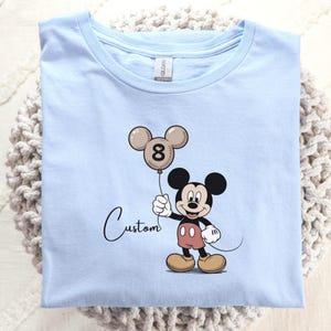 May include: A pale blue t-shirt with a Mickey Mouse cartoon design. Mickey Mouse is holding a balloon shaped like his head, with the number 8 on it. The word "Custom" is written in cursive below the image.