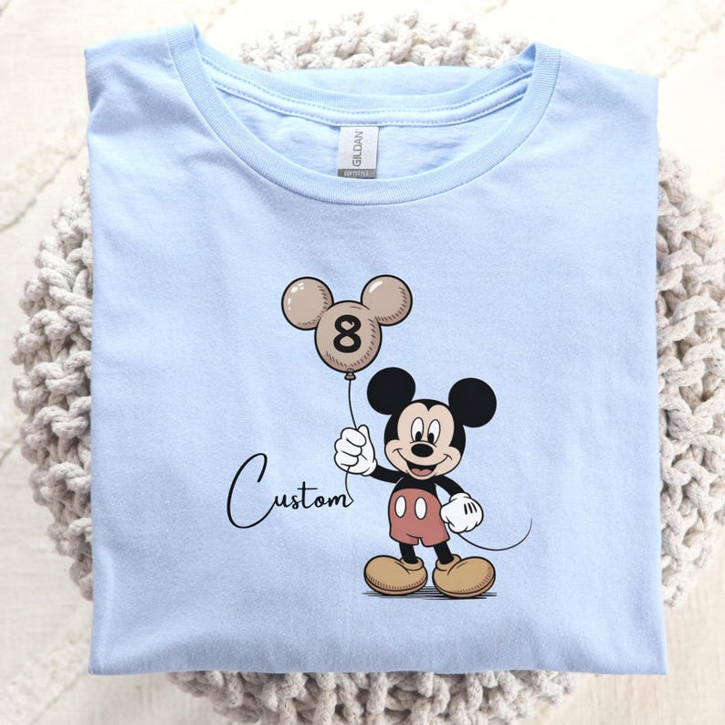 May include: A pale blue t-shirt with a Mickey Mouse cartoon design. Mickey Mouse is holding a balloon shaped like his head, with the number 8 on it. The word "Custom" is written in cursive below the image.