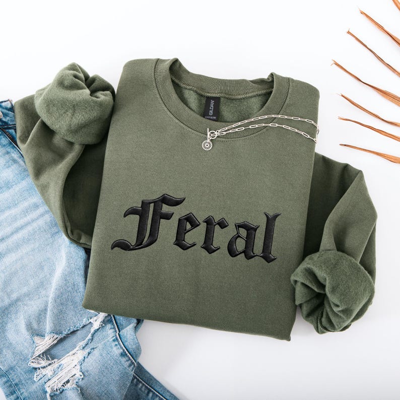 May include: An olive green sweatshirt with the word "Feral" embroidered in black gothic lettering. A silver necklace with a circular pendant rests on the shirt. The sweatshirt is folded over a pair of blue jeans.