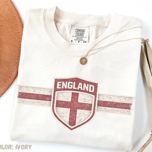 May include: An ivory short-sleeve t-shirt featuring a distressed red shield graphic with the word ENGLAND and a horizontal red stripe. The shirt has a crew neckline and a Comfort Colors brand label inside the collar. A gold-tone necklace is placed on top.