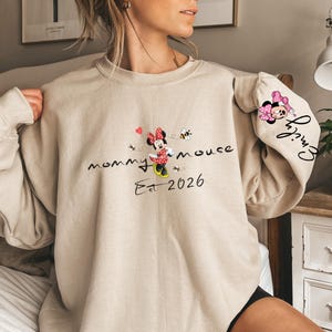 Pode incluir: Camisola bege com um gr&aacute;fico da Minnie Mouse e o texto "mommy mouse Est 2026". A manga tem um gr&aacute;fico da Minnie Mouse e o nome "Emily". O design inclui cora&ccedil;&otilde;es e abelhas.