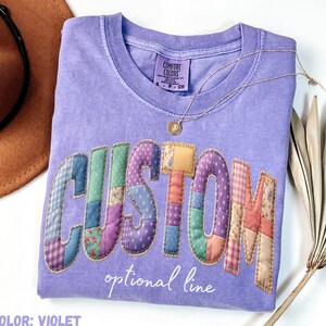 May include: A violet Comfort Colors t-shirt with the word "CUSTOM" in a patchwork design. The shirt has an optional line of text below the word. A gold necklace and a brown hat are also visible.
