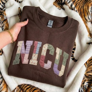 Könnte beinhalten: Ein braunes Sweatshirt mit Rundhalsausschnitt und dem Wort "NICU" auf der Vorderseite in einem Patchwork-Design. Die Patchwork-Buchstaben weisen verschiedene Blumen- und Steppmuster in Rosa, Blau und Grün auf. Das Sweatshirt wird auf einer weißen Decke mit Tigerprint präsentiert.