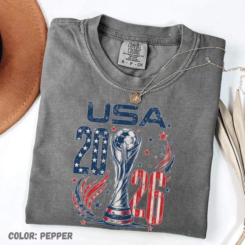 K&ouml;nnte beinhalten: Ein pfefferfarbenes T-Shirt mit einem Grafikdruck, der den Text USA 2026, einen Fu&szlig;ballpokal sowie rote, wei&szlig;e und blaue Sterne und Streifen zeigt. Das T-Shirt hat einen Rundhalsausschnitt und ein Comfort Colors Markenetikett am Kragen.