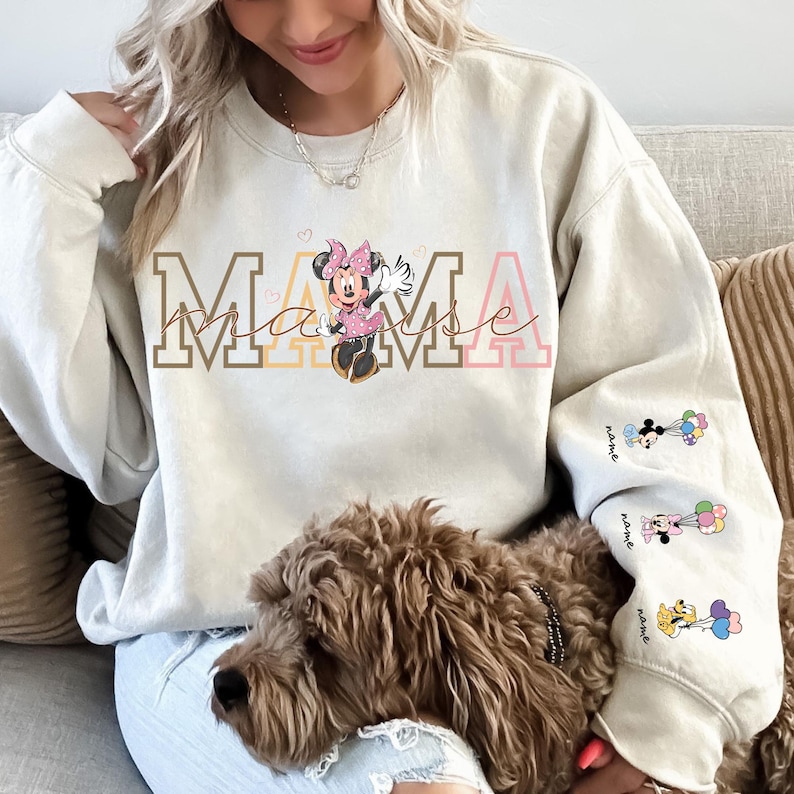May include: Beige sweatshirt with the word "MAMA" in brown, yellow, and pink lettering, featuring a Minnie Mouse graphic. The sleeves have additional Minnie Mouse graphics with balloons. A dog is in the foreground.