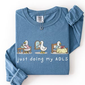 May include: A blue long-sleeve t-shirt featuring a graphic of three ducks performing daily activities with the text "just doing my ADLS" printed below. The shirt has a Comfort Colors brand label at the inner neckline and a silver sun-shaped pendant.