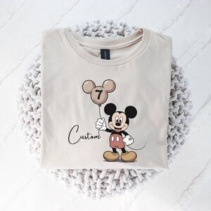 May include: A beige t-shirt with a Mickey Mouse cartoon holding a balloon with the number 7. The word "Custom" is written in cursive below the character. The shirt is folded on a white knitted surface.