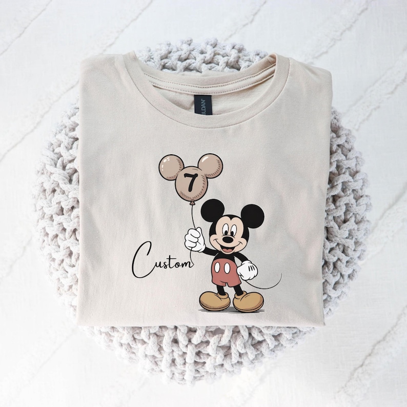 May include: A beige t-shirt with a Mickey Mouse cartoon holding a balloon with the number 7. The word "Custom" is written in cursive below the character. The shirt is folded on a white knitted surface.