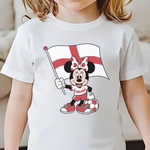 May include: A white short-sleeved t-shirt featuring a graphic of Minnie Mouse holding an England flag. Minnie wears a red and white outfit with a matching bow, and stands next to a soccer ball. The shirt includes the text ENGLAND on her top.