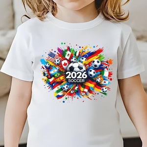 May include: White t-shirt featuring a colorful soccer-themed graphic. The design includes soccer balls, flags from various countries, and the text "2026 SOCCER". The graphic is a vibrant mix of colors, with a central soccer ball.