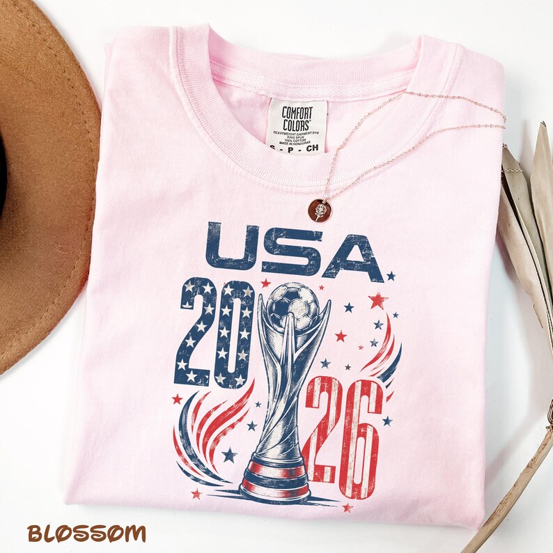 K&ouml;nnte beinhalten: Ein hellrosa T-Shirt mit einem Aufdruck, der den Text USA 2026, einen Fu&szlig;ballpokal sowie rote, wei&szlig;e und blaue Sterne und Schw&uuml;nge zeigt. Das T-Shirt hat ein Comfort Colors Markenetikett am inneren Halsausschnitt und eine d&uuml;nne Goldkette.