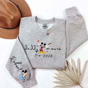 Pode incluir: Um moletom cinza com o texto "Daddy Mouse Est 2026" e um gr&aacute;fico do Mickey Mouse. A manga apresenta o nome "Robert" e um gr&aacute;fico do Mickey Mouse. O moletom &eacute; de gola redonda.