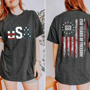 May include: A dark charcoal gray t-shirt featuring a distressed USA graphic on the front with red, white, and blue flag patterns. The back displays a distressed American flag design with a circle of stars, the years 1776-2026, and 250 YEARS OF FREEDOM.