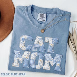 May include: A blue jean Comfort Colors t-shirt with the words "CAT MOM" appliqued in a patchwork design of floral and gingham patterns. A gold necklace with a pendant rests on the shirt. The shirt is a size small.